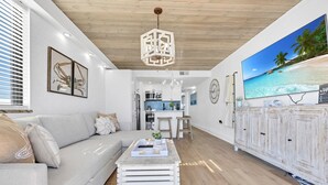 En moderne stue med sofa, sofabord og stort fjernsyn, der viser et tropisk strandmotiv.