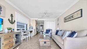 A spacious living room with a beige sofa, a coffee table, and a dining area with a white table and chairs. There is a kitchen in the background with white cabinets and a ceiling fan. - Direct Ocean Front w/washer and dryer! (Myrtle Beach)