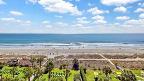 A beach with a clear sky, a sandy shore, and a grassy area with palm trees and sun loungers. - Direct Ocean Front w/washer and dryer! (Myrtle Beach)