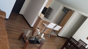 Ein modernes Wohnzimmer mit braunem Sofa, einem Glastisch mit gewebtem Korb, einer weißen Lampe und einer Küchenzeile mit Kühlschrank und Holzschränken.