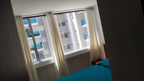 Ein Zimmer mit großem Fenster, Vorhängen und einem Bett mit blauer Bettdecke.