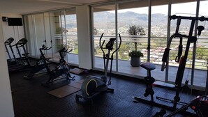 Ein Fitnessraum mit Ergometer, Rudergerät und Laufband. Große Fenster bieten Blick auf die Berge.