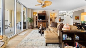A spacious living room with a large window, a ceiling fan, a sofa, a coffee table, and a bookshelf. - Beach Colony East 8B (Pensacola)