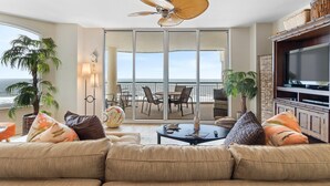 A spacious living room with a large sofa, a ceiling fan, and a television. There is a balcony with a view of the ocean. - Beach Colony East 8B (Pensacola)