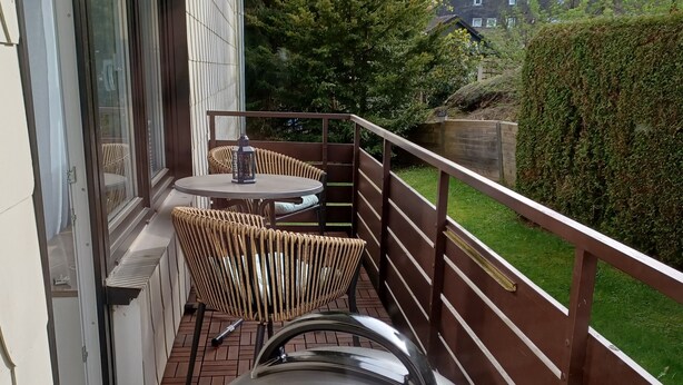 Ein Balkon mit Korbmöbeln, einem Grill und Blick auf einen Garten sowie Gebäude.