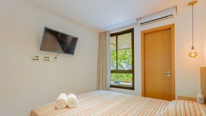 Hotellihuone, jossa on sänky, seinälle asennettu televisio, ilmastointilaite, ikkuna verhoilla ja yöpöytälamppu.