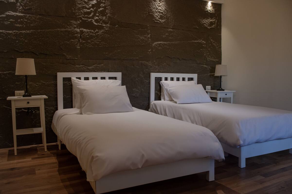 A room with two beds, each with white bedding and headboards, against a textured wall. There are bedside tables with lamps on either side of the beds.