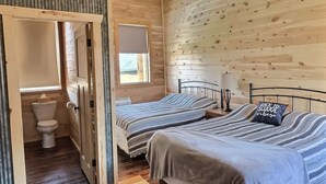Une chambre avec deux lits, un ventilateur de plafond, des murs en bois, un plancher en bois, une fenêtre et une porte donnant sur une salle de bain.