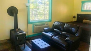 A living room with a black leather sofa, a small table, a black wood-burning stove, and a window with a view of trees. - Cabin Getaway (Cashmere)