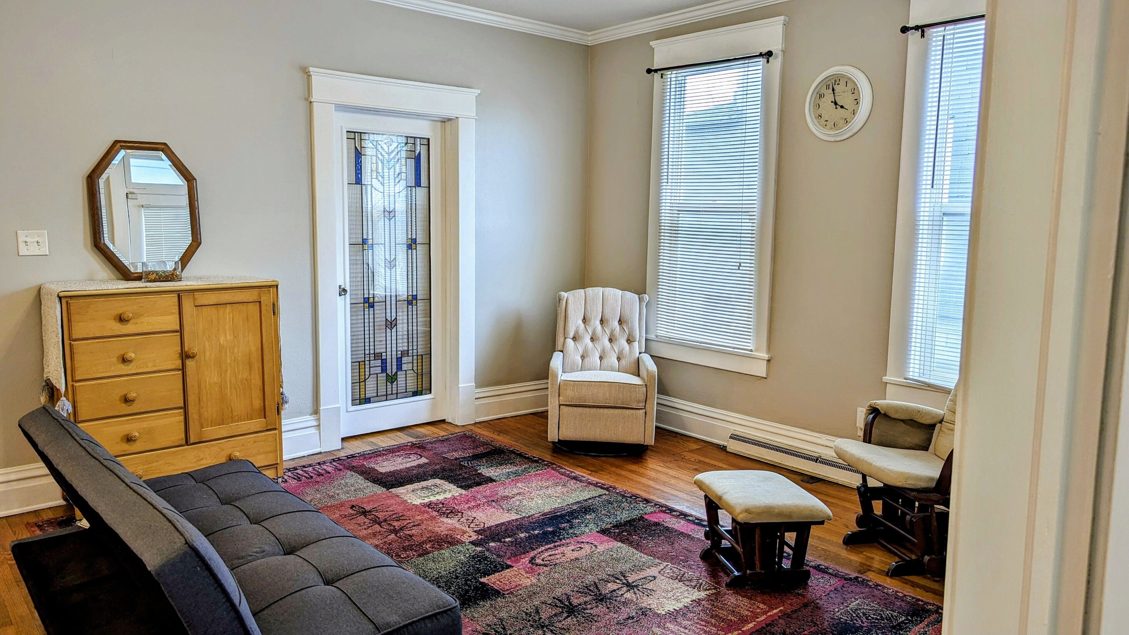 A room with a wooden dresser, a tufted armchair, a bench, a colorful rug, and a large mirror.