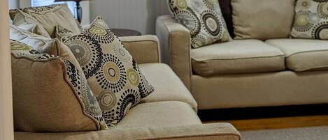 Un salon avec deux canapés beiges, des coussins à motifs, un lampadaire rouge et de grandes fenêtres équipées de stores.