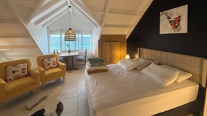 A bedroom with a bed, two yellow armchairs, a dining table, and a view of the sea.