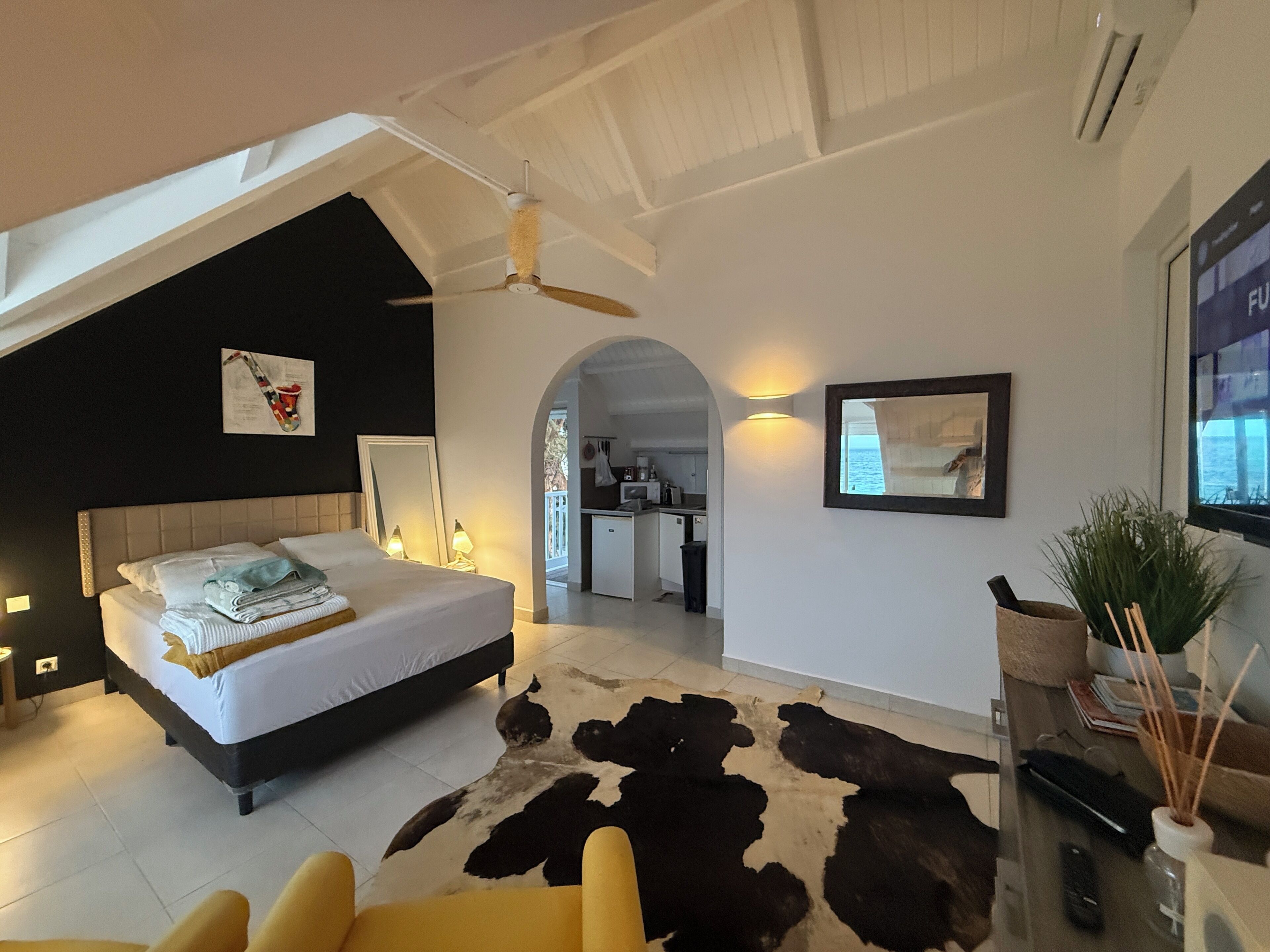 Een slaapkamer met een bed, een plafondventilator, een kleden van koeienhuid, een gele fauteuil en uitzicht op de keuken en het balkon.