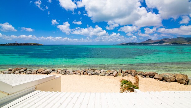 A beach view with clear turquoise water, a rocky shoreline, and a white building with a sloped roof.