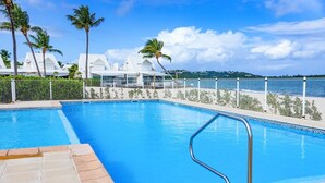 A rectangular swimming pool with a metal ladder, surrounded by a fence and palm trees, with a view of a white building in the background.
