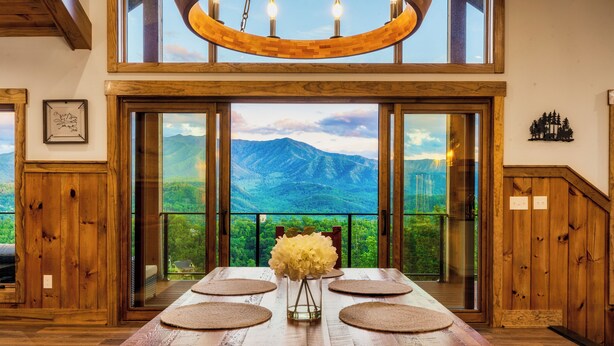 Une table à manger avec des sets de table, un vase rempli de fleurs et une vue sur les montagnes par de grandes fenêtres.