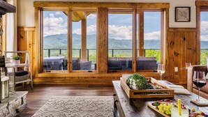 Une pièce chaleureuse aux murs en bois et aux grandes fenêtres donnant sur les montagnes. On y trouve un coin salon avec une table et un plateau de raisins.