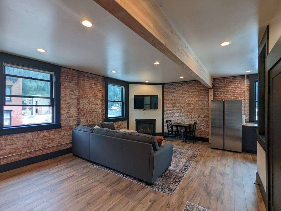 A modern living room with a brick wall, a gray sofa, a fireplace, and a kitchen area with a refrigerator.