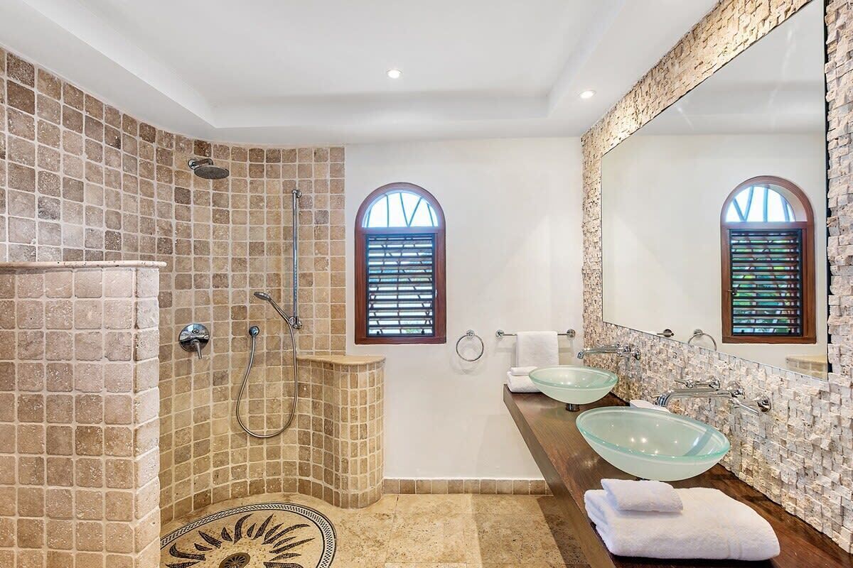 A bathroom with a stone-tiled shower, two arched windows, a glass bowl sink, and a wooden vanity.