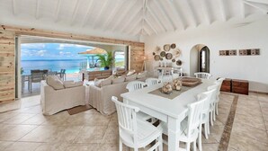 A spacious living area with a large dining table, white chairs, a sofa set, and a view of the ocean through a glass door. - Villa Happy Bay – Secluded Creole Elegance near Grand Case, St. Martin (Saint Martin)
