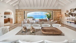 A spacious living area with a large window overlooking a beach, white furniture, and a wooden wall. - Villa Happy Bay – Secluded Creole Elegance near Grand Case, St. Martin (Saint Martin)