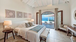 A bedroom with a bed, bedside tables, a bench, and a view of the ocean through an open door. - Villa Happy Bay – Secluded Creole Elegance near Grand Case, St. Martin (Saint Martin)