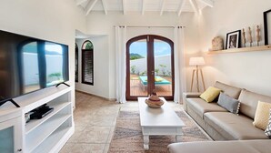 A modern living room with a TV, white furniture, a coffee table, and a view through a glass door to an outdoor area. - Villa Happy Bay – Secluded Creole Elegance near Grand Case, St. Martin (Saint Martin)