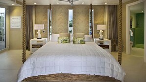 A large bed with a white quilted bedspread, two nightstands with lamps, a ceiling fan, and a view of the outdoors through large windows. - Villa C’est La Vie – Luxury Beachfront Villa on Plum Bay, St. Martin with Private Chef (Marigot)