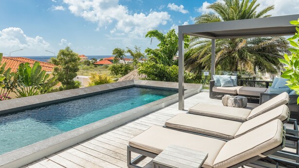 A swimming pool with a wooden deck and lounge chairs, surrounded by tropical plants and a view of houses. - The Pearl of Jan Thiel 1 – Luxury Designer Villa with Private Pool & Resort Facilities in Curaçao (Willemstad)