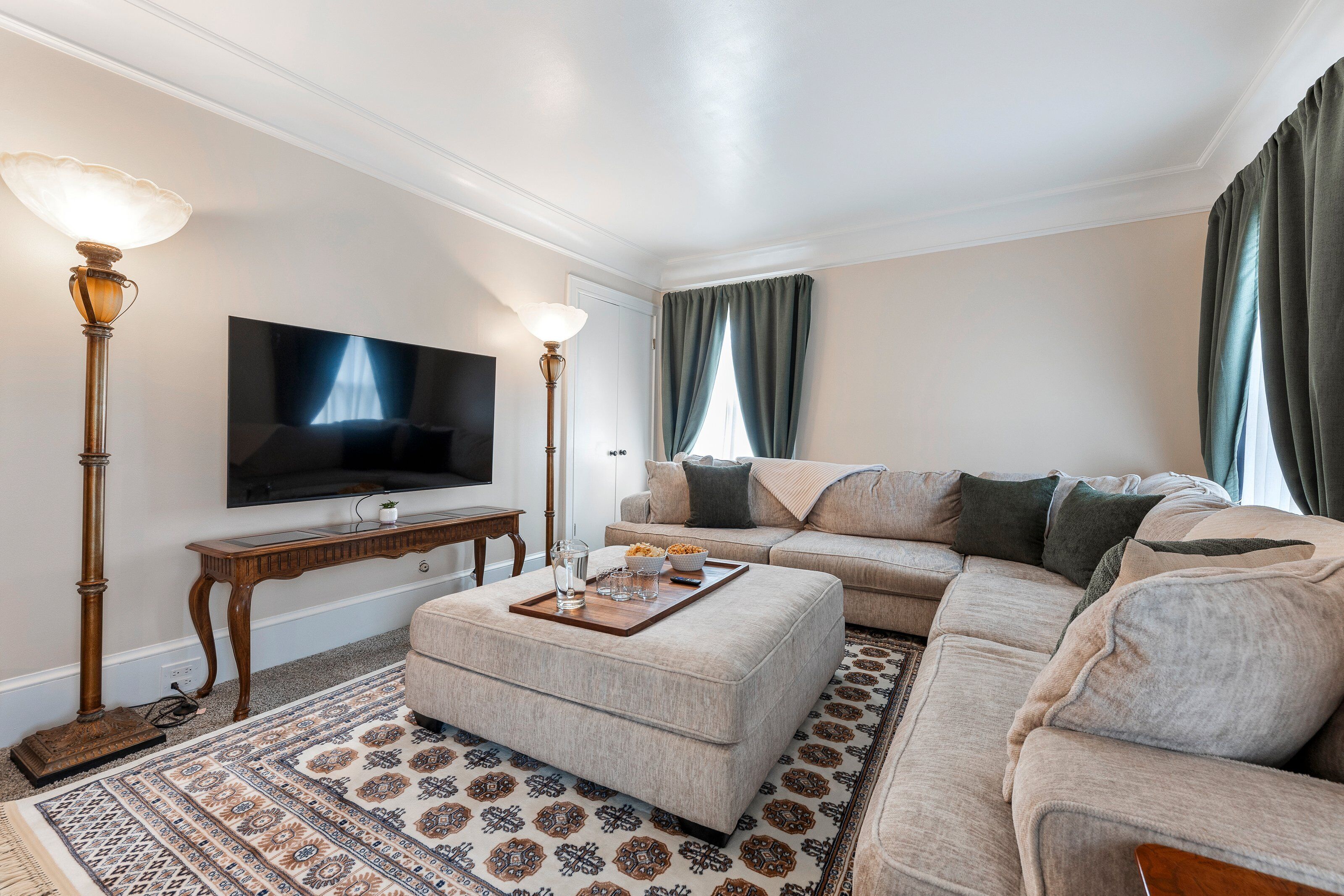 Ein Wohnzimmer mit einem Ecksofa, einem Flachbildfernseher, einem Couchtisch mit Tablett und einem gemusterten Teppich.