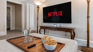 A living room with a TV displaying "NETFLIX," a wooden coffee table with snacks, glasses, and a remote control. - - Beautiful Condo - King Bed - Free Waffle Breakfast - (Geneva)