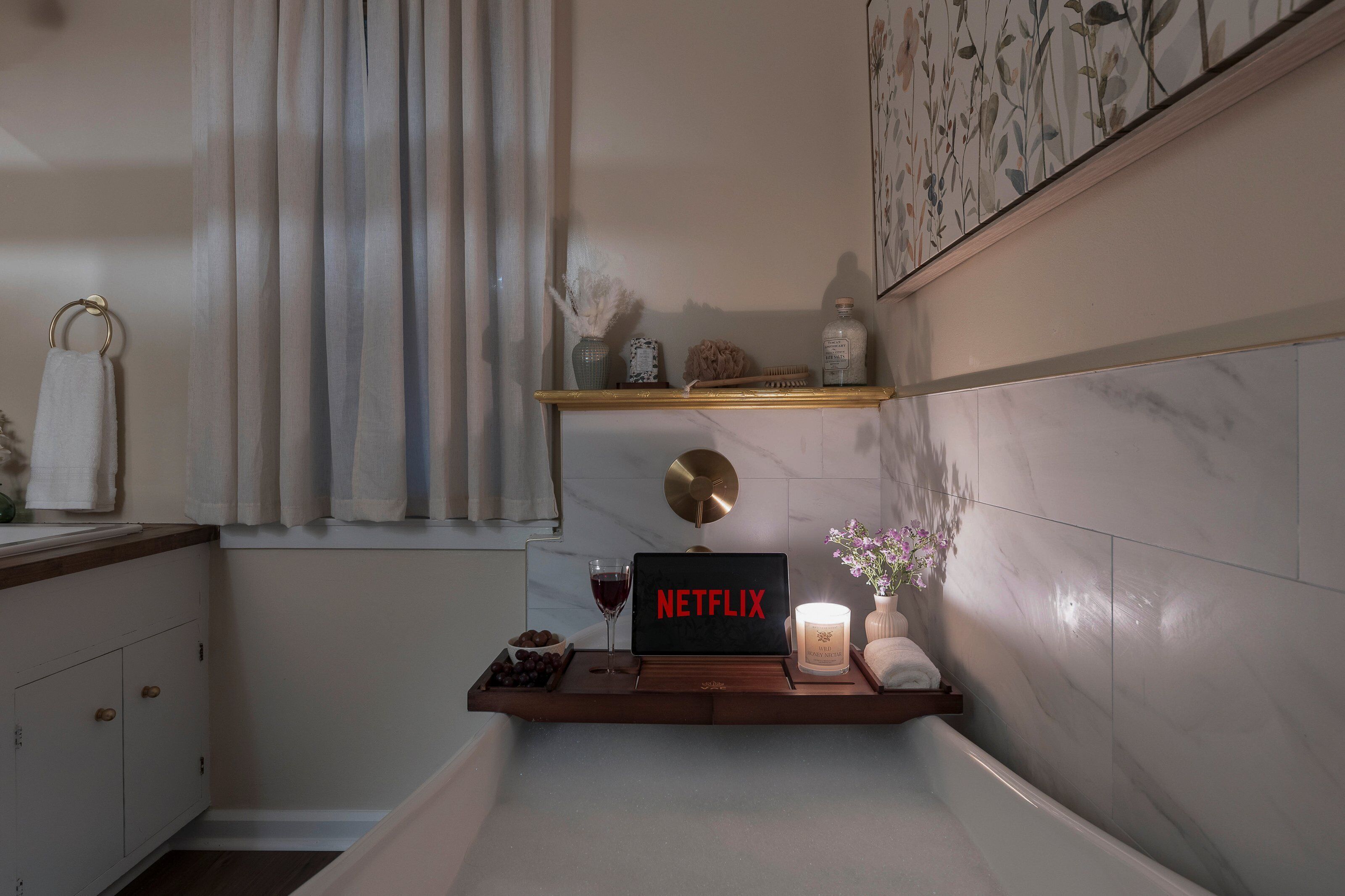 A bathroom with a wooden shelf holding a Netflix sign, a candle, a glass of wine, and a small bowl of grapes.