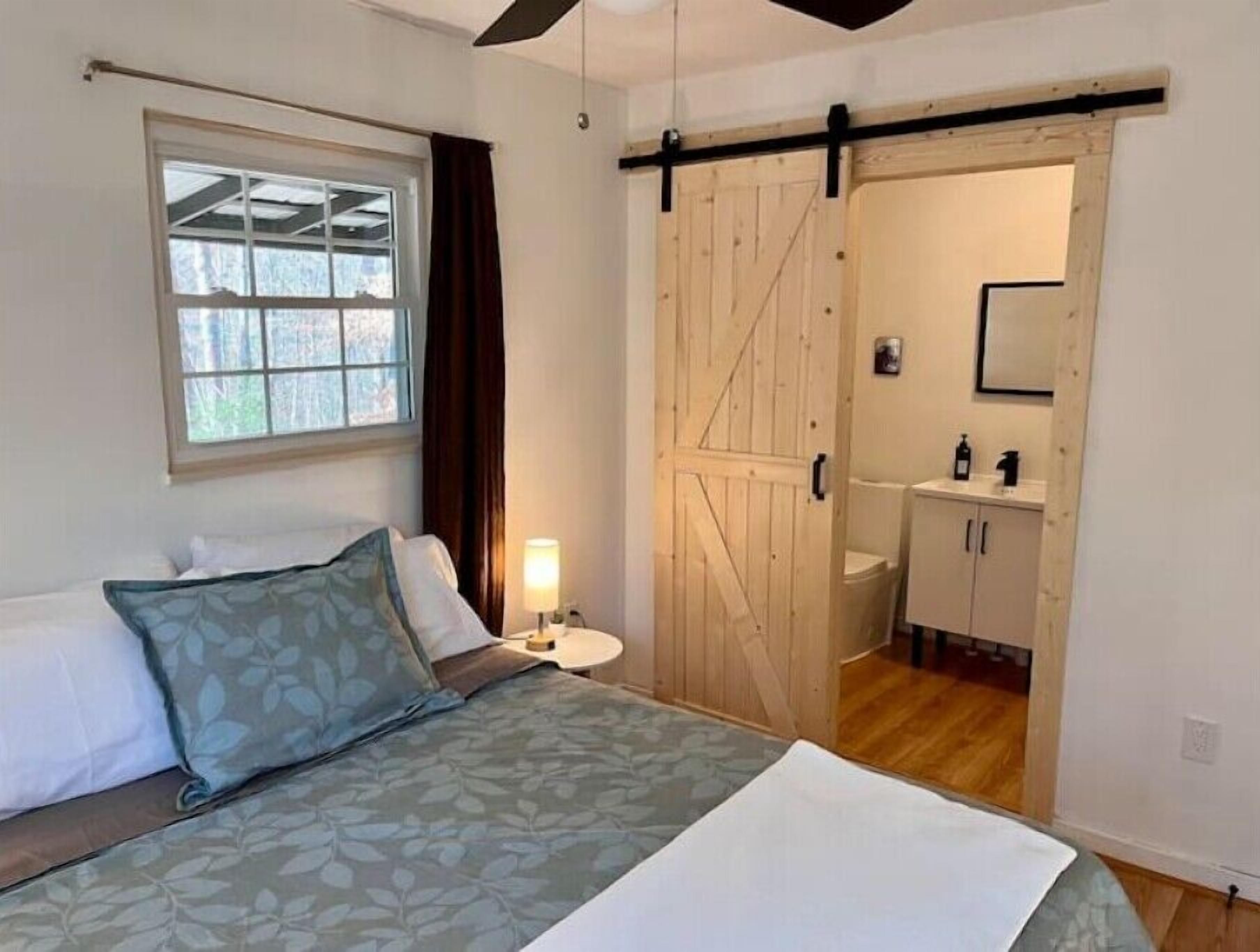 Une chambre à coucher comprenant un lit, une fenêtre, une table de chevet, une lampe, une porte coulissante de style rustique et une salle de bain avec un lavabo et des placards.