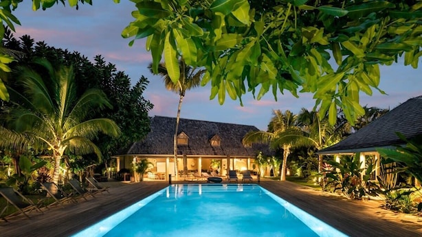Une piscine entourée de chaises longues et de plantes tropicales, avec un bâtiment en arrière-plan.