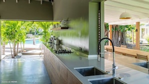 Une cuisine moderne dotée d’un grand îlot central, d’appareils électroménagers intégrés et offrant une vue sur une piscine et un espace verdoyant.