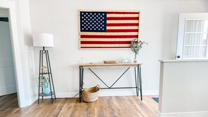 Une pièce avec un plancher en bois, un drapeau américain accroché au mur, un lampadaire, une petite table avec un vase et un panier en osier.