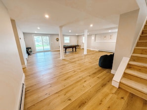 Une pièce spacieuse avec un plancher en bois, une table de billard, un escalier et une grande fenêtre.