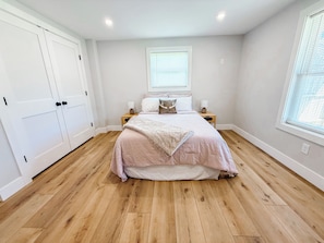 Een slaapkamer met een bed, twee nachtkastjes, een raam met jaloezieën en een deur.