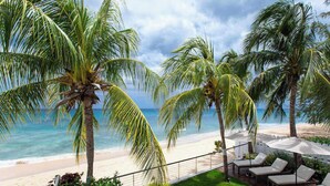 Vista de una playa con palmeras, una orilla arenosa y sillones de playa con sombrillas.