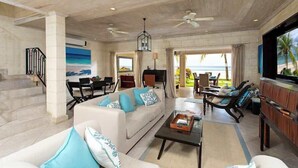 Un amplio salón con un sofá blanco, una mesa de centro y un televisor. Hay varias sillas y una mesa de comedor. La habitación tiene techo de madera y grandes ventanales con vistas a la playa.