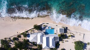 Vue aérienne d’une propriété située en bord de mer, avec une piscine, plusieurs bâtiments et une plage de sable.