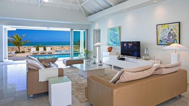 Un salon moderne doté d’un grand téléviseur, de sièges confortables, d’une vue sur la piscine et d’une terrasse donnant sur la plage.