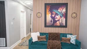 Un sofá color turquesa con estampado de leopardo y cojines blancos, un cuadro abstracto enmarcado, y dos luces fijadas a la pared.