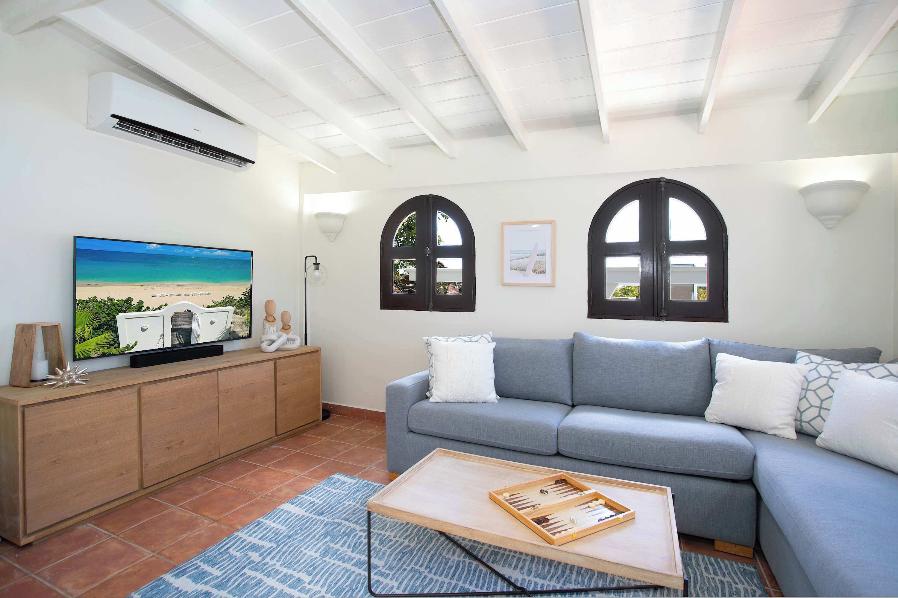 A living room with a grey sofa, a wooden coffee table with a board game, a flat-screen TV on a wooden cabinet, and two arched windows with pictures.