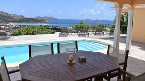A poolside area with a table and chairs, a glass candle holder, and a view of the sea and distant hills.