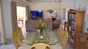 A living room with a glass-top dining table surrounded by wicker chairs, a sofa, a television, and a bookshelf.