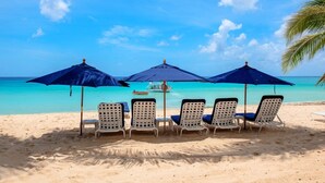 A beach with four lounge chairs under blue umbrellas, a boat in the water, and clear turquoise sea and sky.