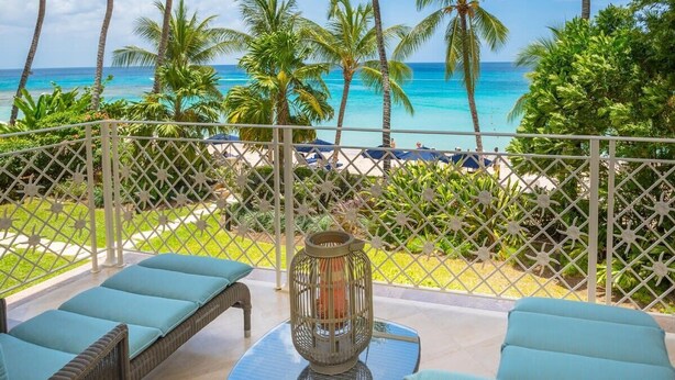 A balcony with wicker furniture and a decorative lantern, overlooking a beach with palm trees and a clear blue ocean.