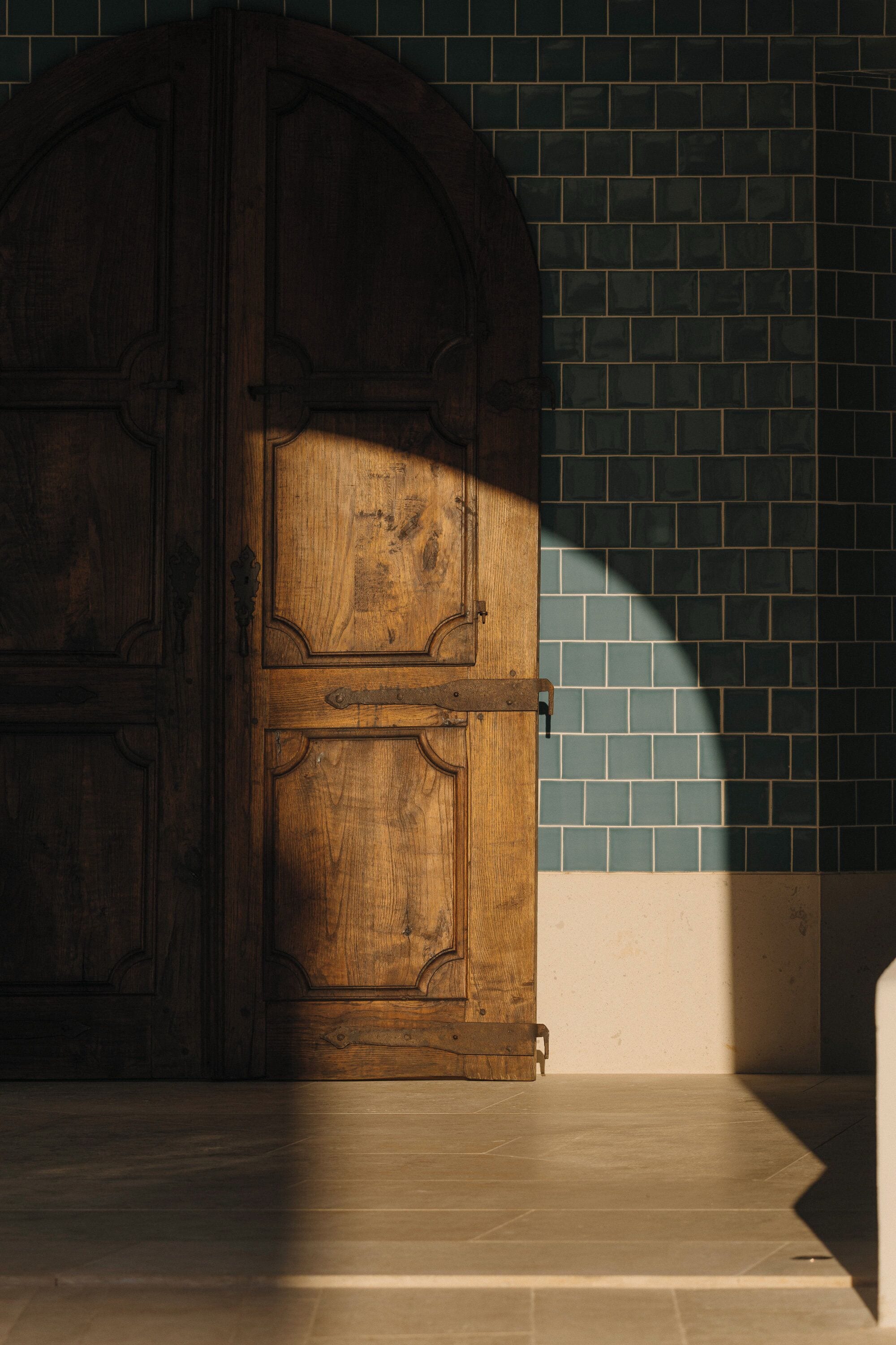 A wooden door with a metal latch, a tiled wall, and a shadow cast on the floor.