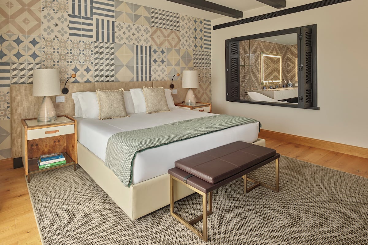 Une chambre d’hôtel moderne avec un grand lit, deux tables de chevet avec des lampes, un banc, un tapis à motifs et un mur décoratif orné de motifs géométriques.